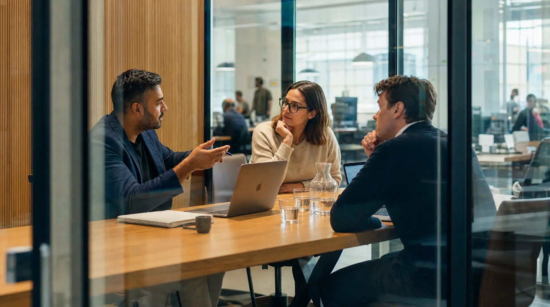 Professionals in a modern office conference room discuss during talent acquisition, with one interviewer gesturing while another candidate thinks and a laptop sits on the table.