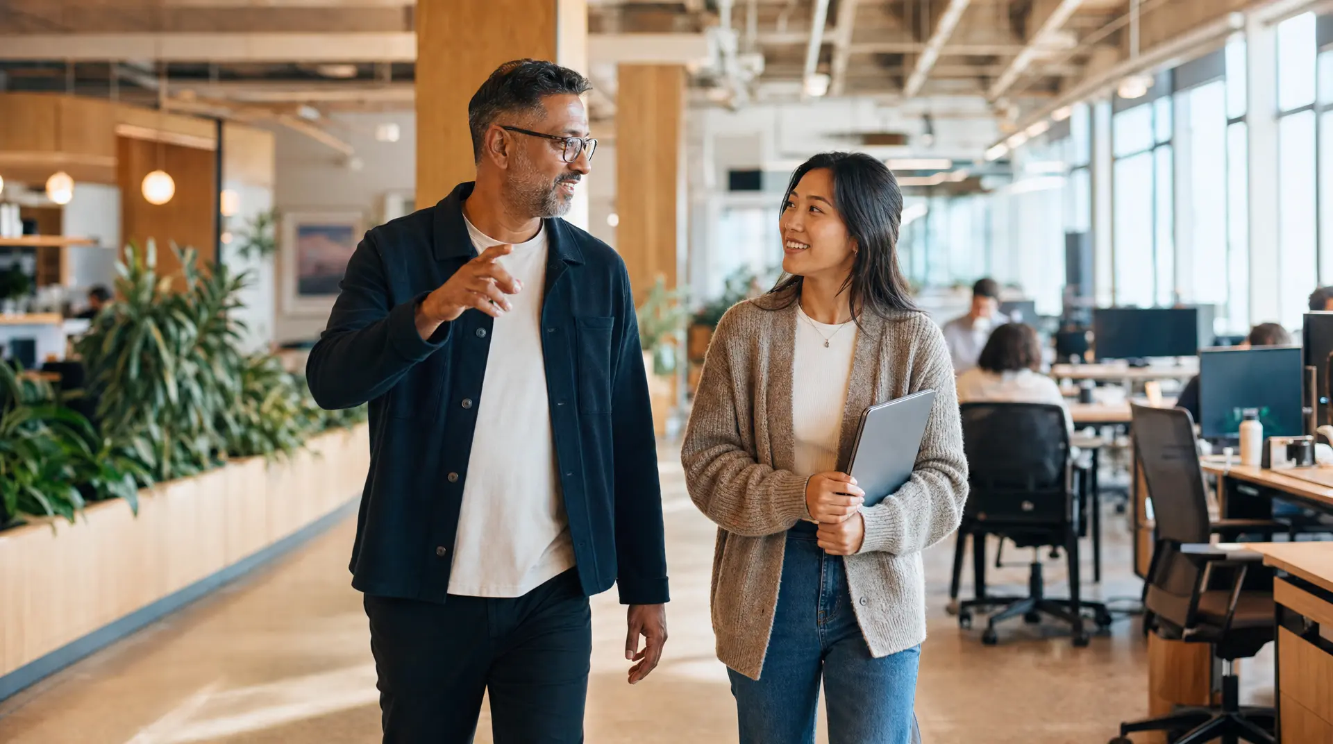 Two colleagues walk through a bright office space, discussing talent acquisition and employer branding while one holds a tablet.