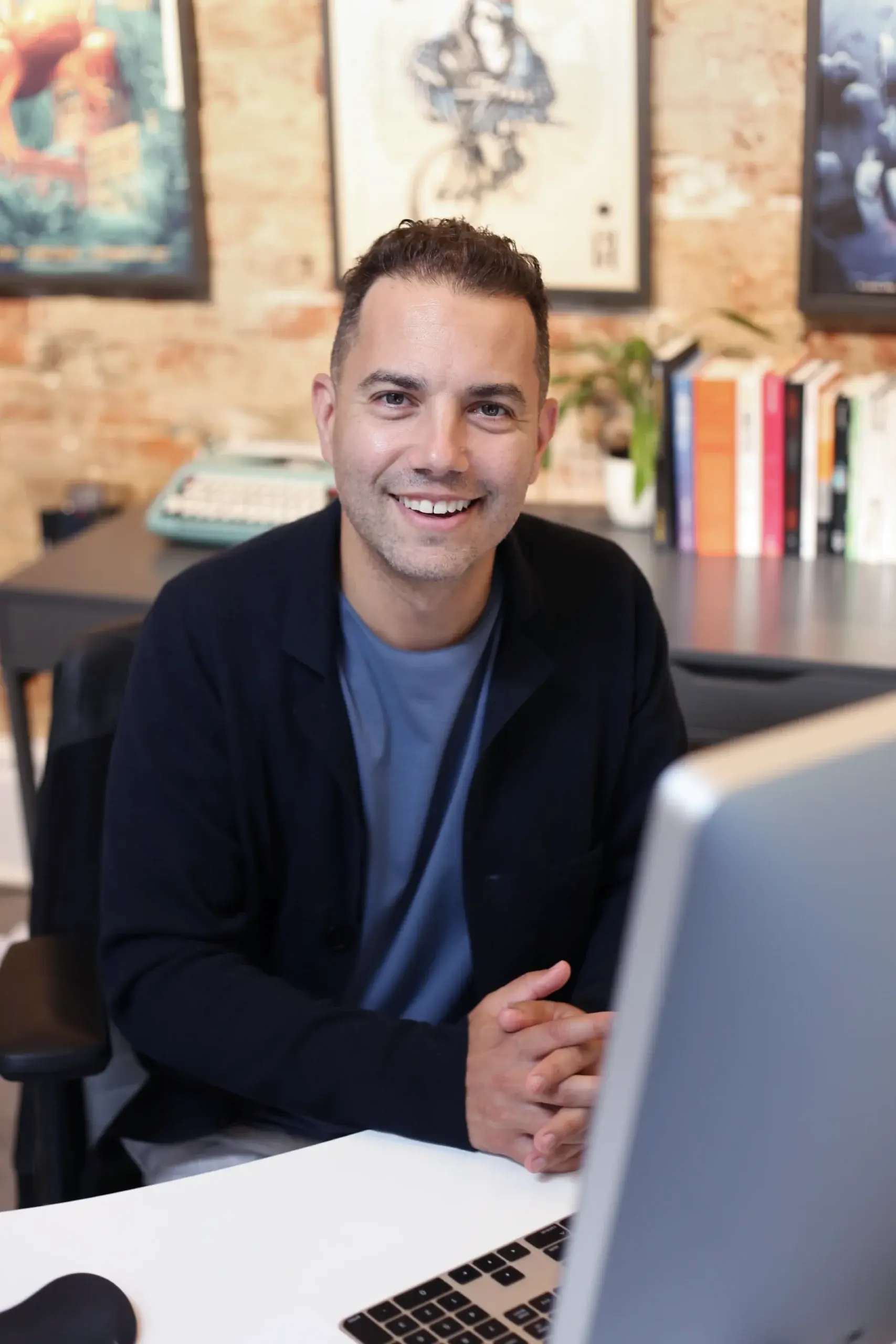 Smiling recruiter sits at a desk with a laptop in a brick-walled office, supporting talent acquisition and recruitment software workflows.