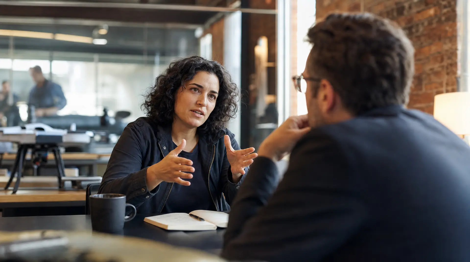 Curly-haired recruiter and interviewee sit across a table with hands gesturing in an open office, focused on improve candidate experience.