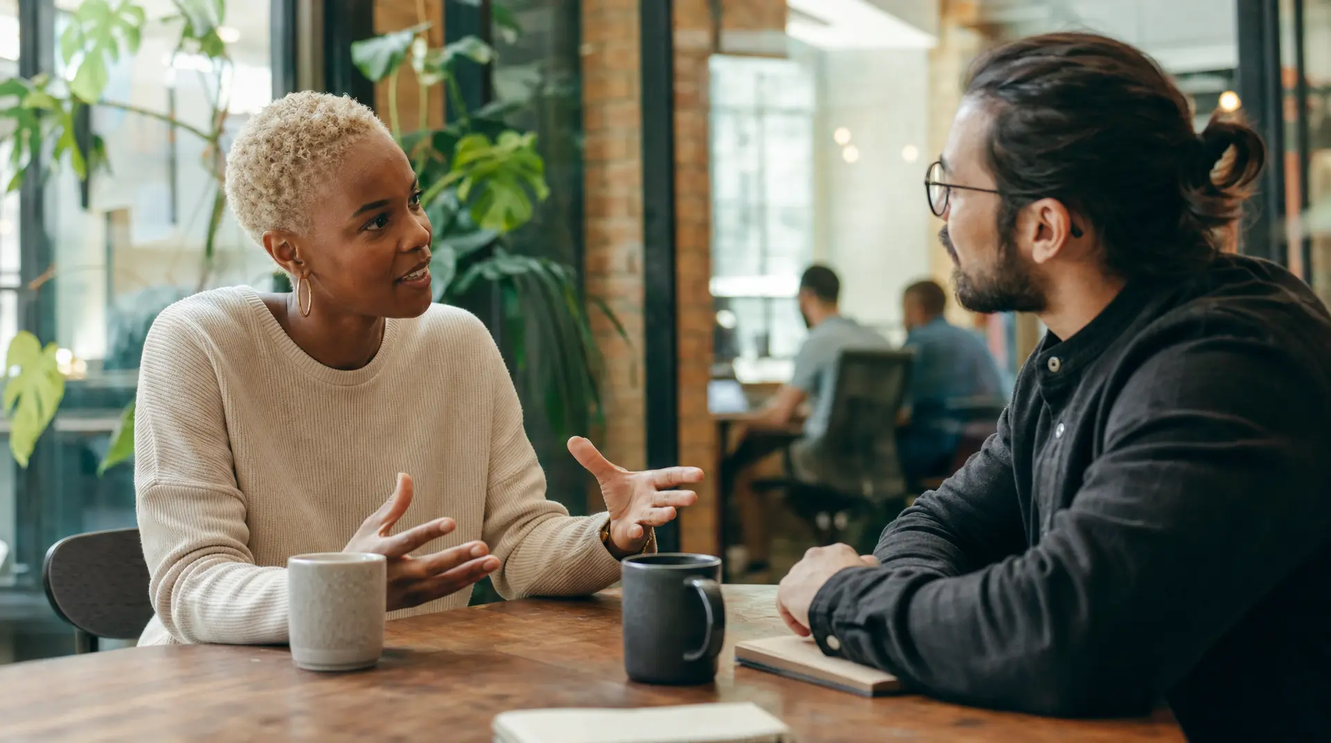 Two professionals sit across from each other at a wooden table in a modern office, gesturing as they talk. The discussion supports improve candidate experience.