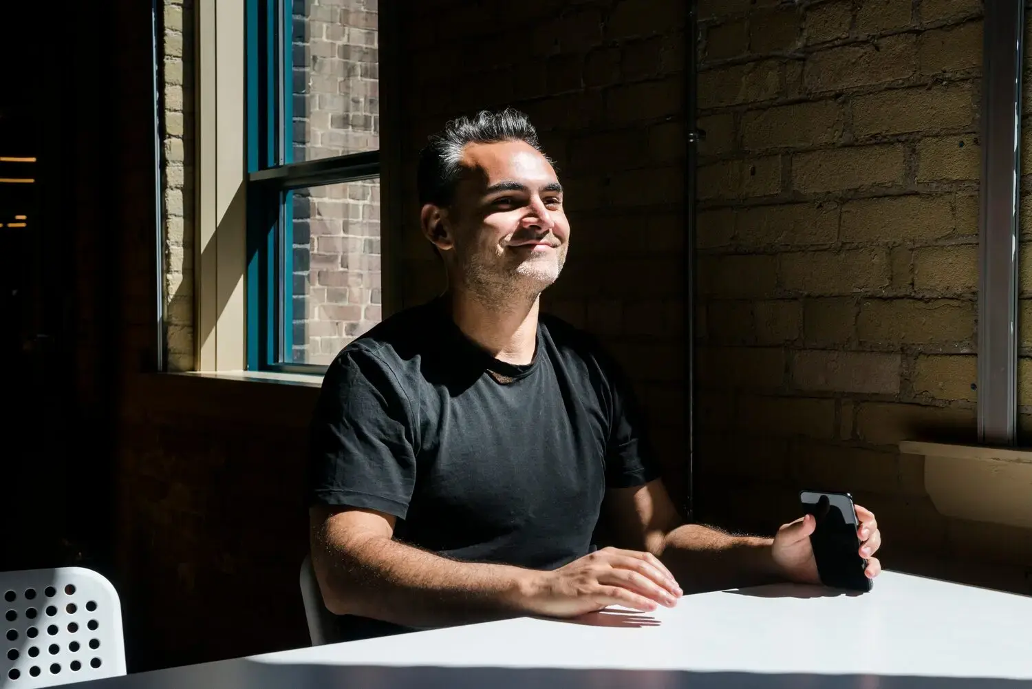 Smiling man seated at a table, holding a smartphone and talking with someone off-camera, supporting improve candidate experience during recruitment.