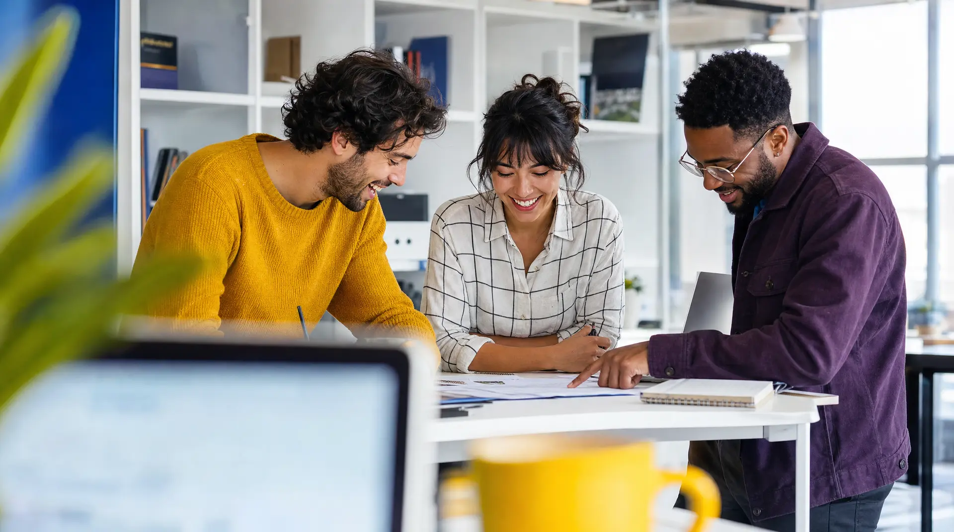 Three colleagues collaborate at a desk with a laptop and paper plans, smiling while one person points during a recruitment process discussion, supporting an improved candidate experience.