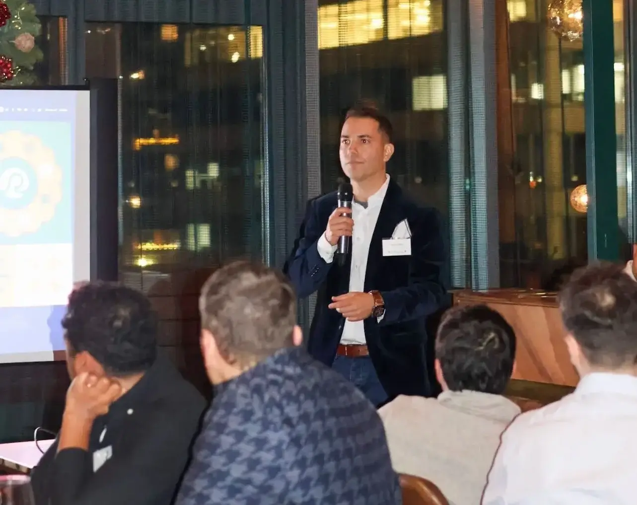 A suited speaker holds a microphone in a conference room while addressing seated attendees in front of a projected slide, as a recruitment webinar session unfolds.