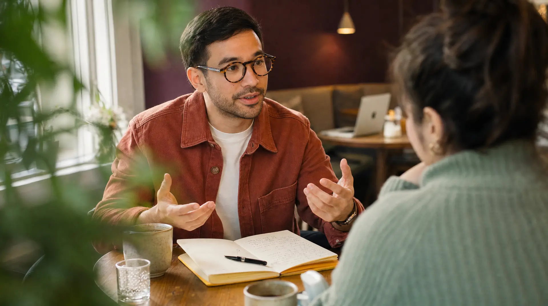 Two professionals meet at a table with notebooks and a laptop during a recruitment process discussion, as the seated man gestures while speaking to the interviewer in a warm office setting.