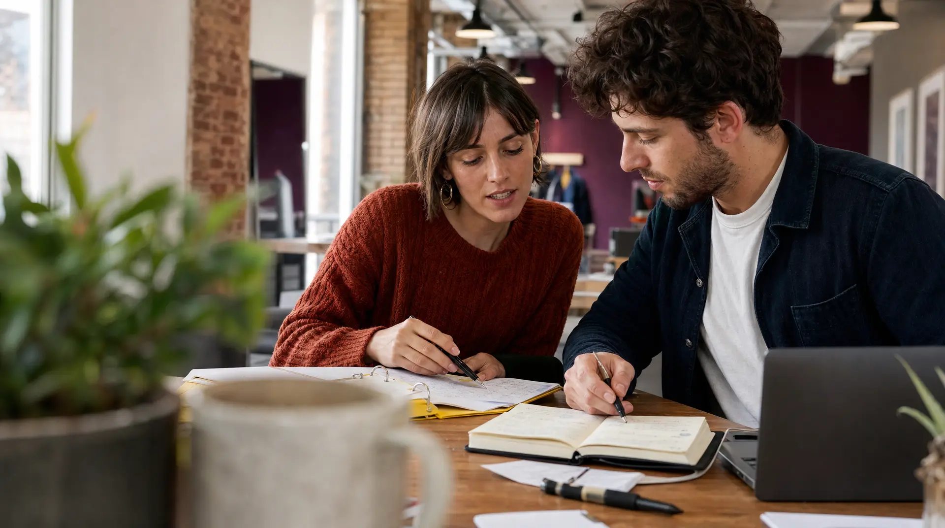Two colleagues sit at a desk in a bright office reviewing paperwork and writing notes on an open folder, collaborating closely on planning tasks.