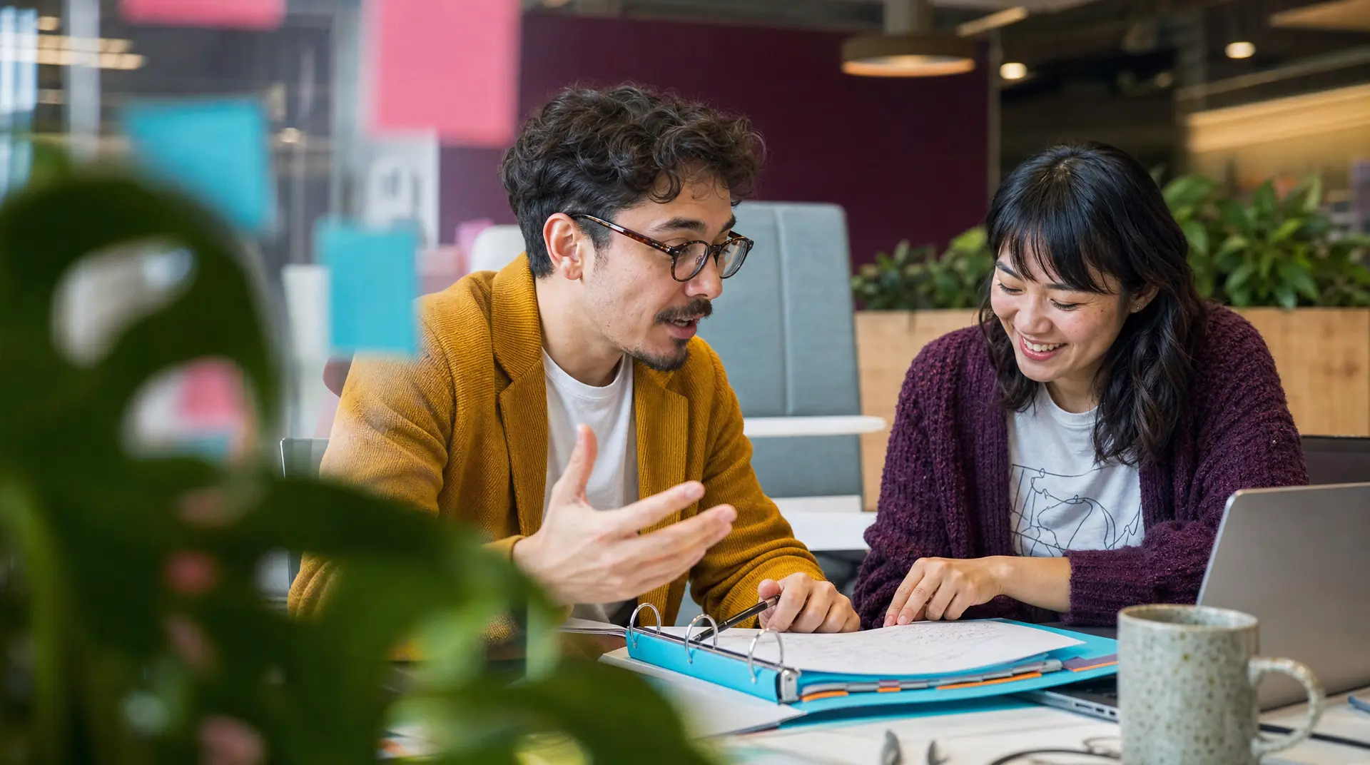 Two professionals sit at a table reviewing notes on a laptop during a candid discussion in a modern office, reflecting talent acquisition teamwork.