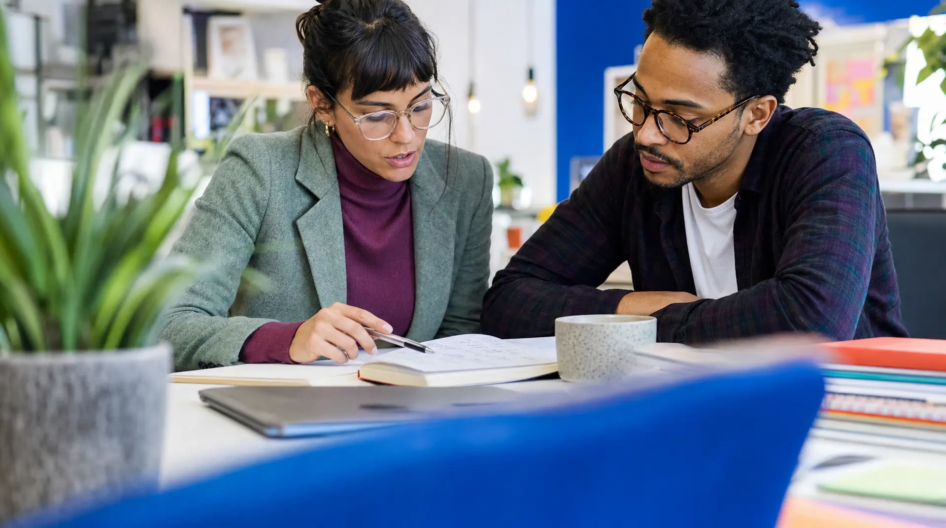 Two colleagues in an office review notes and a document on a table with a notebook and coffee, reflecting the recruitment process planning and coordination.