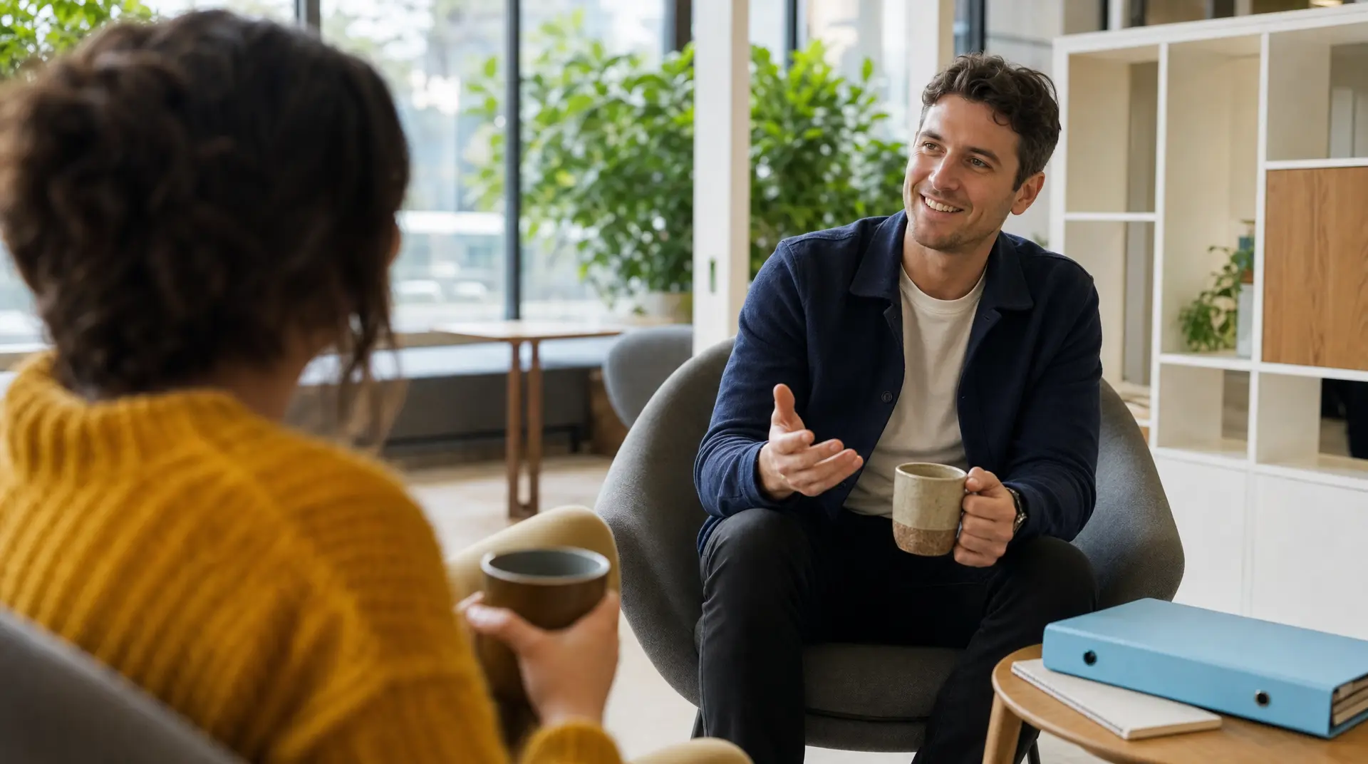 In an office lounge, a recruitment consultant smiles while speaking in an interview, holding a coffee mug across from a seated candidate during candidate recruitment.