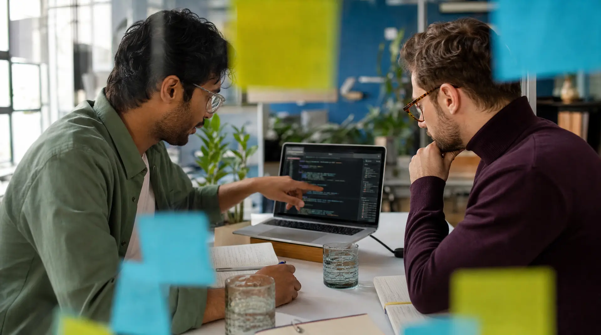 Two colleagues review code on a laptop at a shared desk in a modern office, collaborating on AI for recruitment workflows.