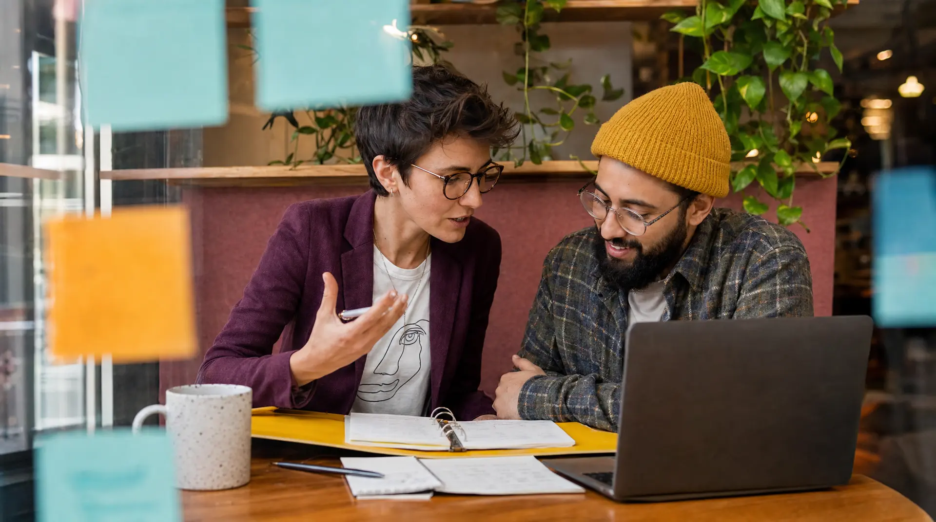 Two colleagues collaborate at a laptop table during a talent acquisition strategy discussion, with notebooks and a coffee mug on the table.