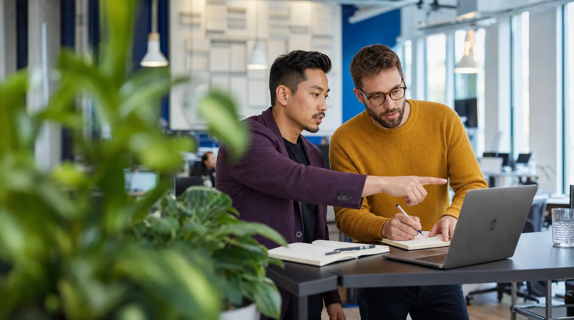 Two colleagues review notes and discuss laptop screens at a shared desk, supporting data analytics in recruitment for smarter hiring decisions.
