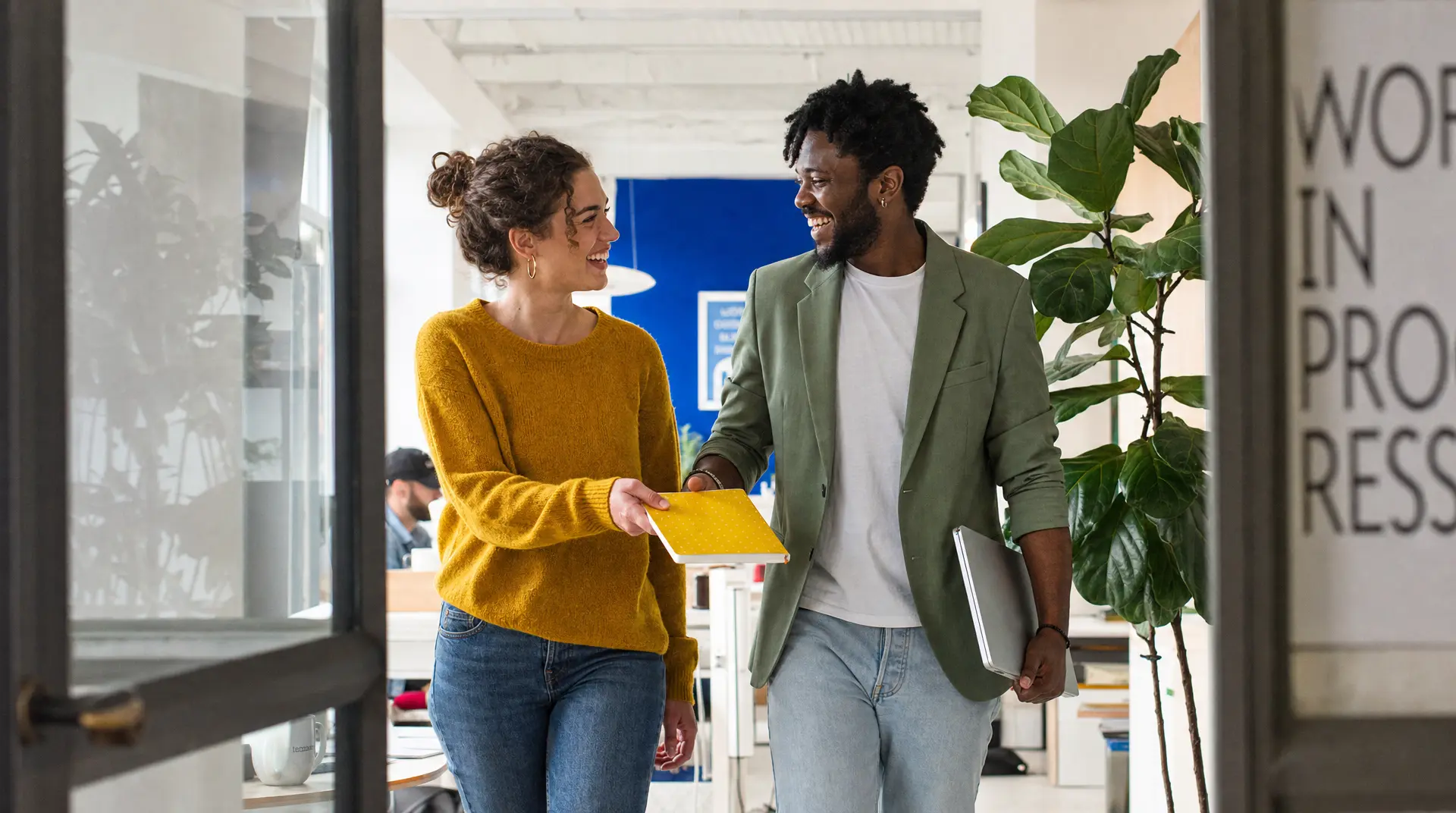 Two coworkers in a bright office corridor smile while handing notes, supporting employee recruitment and improve candidate experience during onboarding discussions.