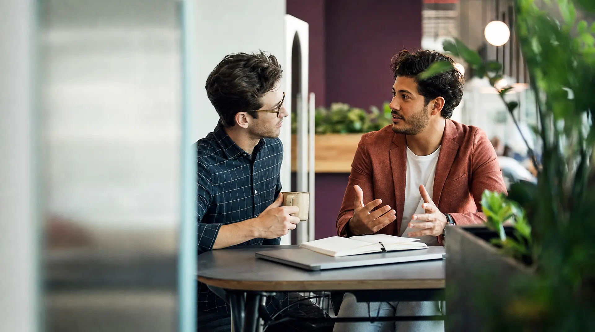 Two professionals sit at a table in a bright office, speaking during a recruitment interview as one holds a coffee mug and a notebook lies open nearby - part of talent acquisition.