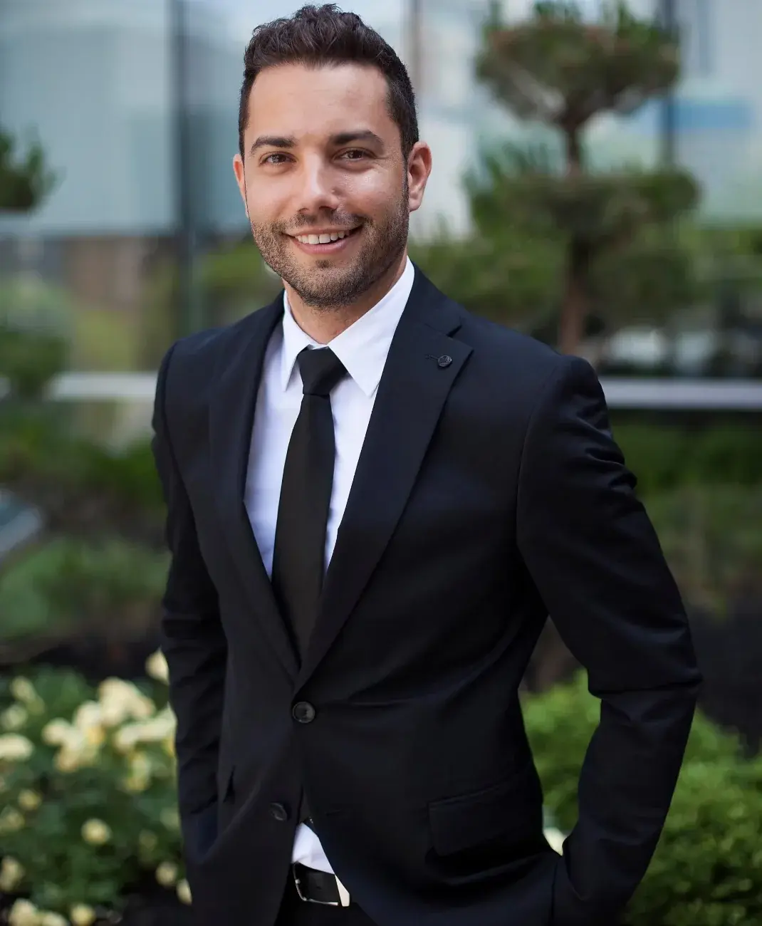 Smiling man in a dark business suit with a black tie standing outdoors near modern office buildings, representing executive search outreach.