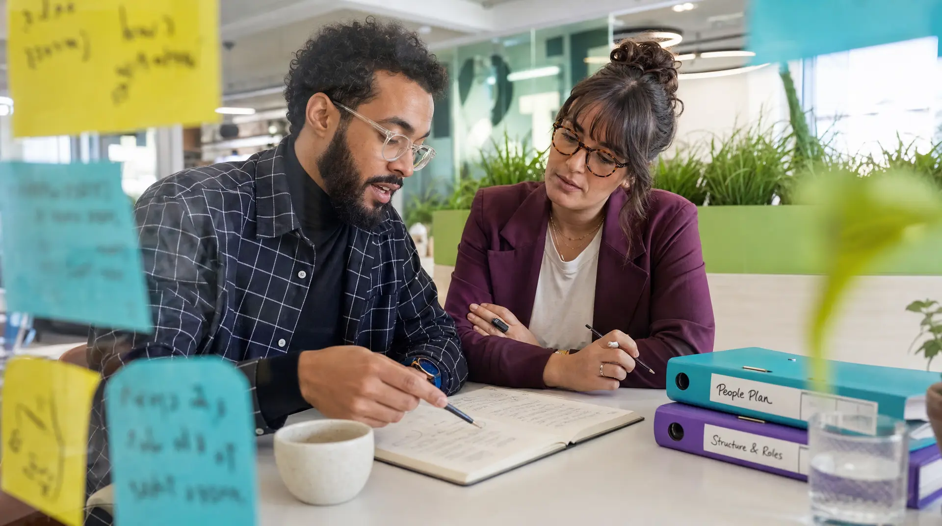 Two colleagues seated at a desk reviewing a recruitment people plan binder labeled "People Plan" and "Structure & Roles," writing notes as part of assessment tools for recruitment.