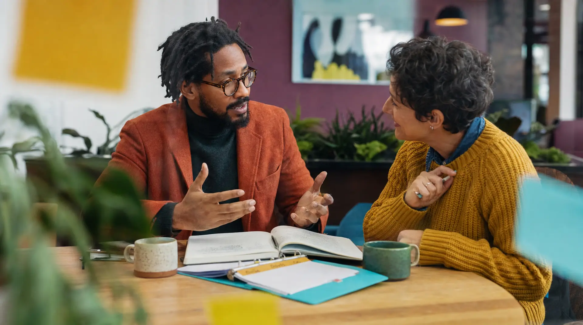 Two professionals discuss an open notebook at a cafe-style table with mugs and colorful folders, reflecting a recruitment process conversation.