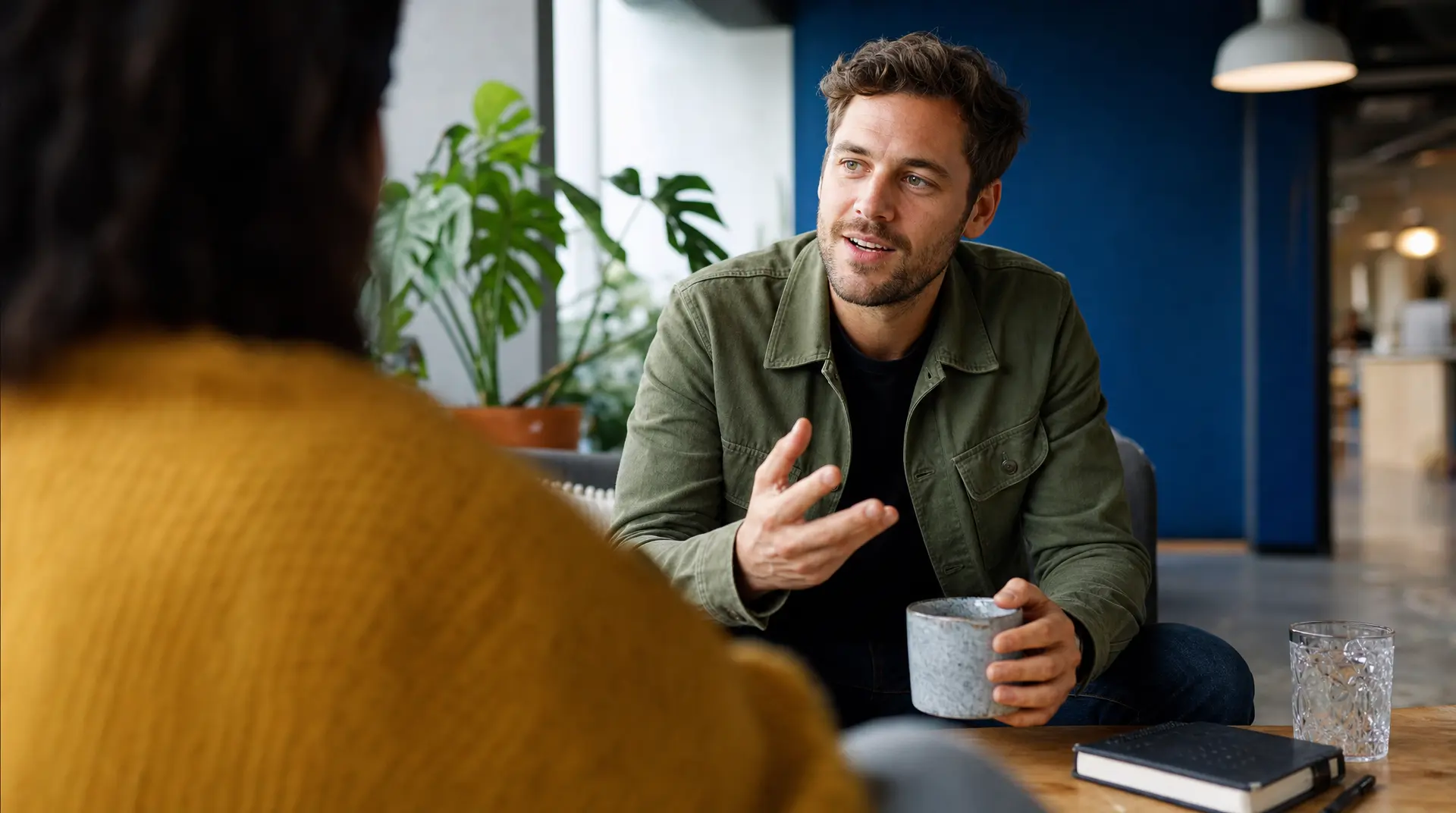 Office interview conversation for talent acquisition recruitment process, interviewer gestures across a meeting table while a candidate listens with a notebook nearby.