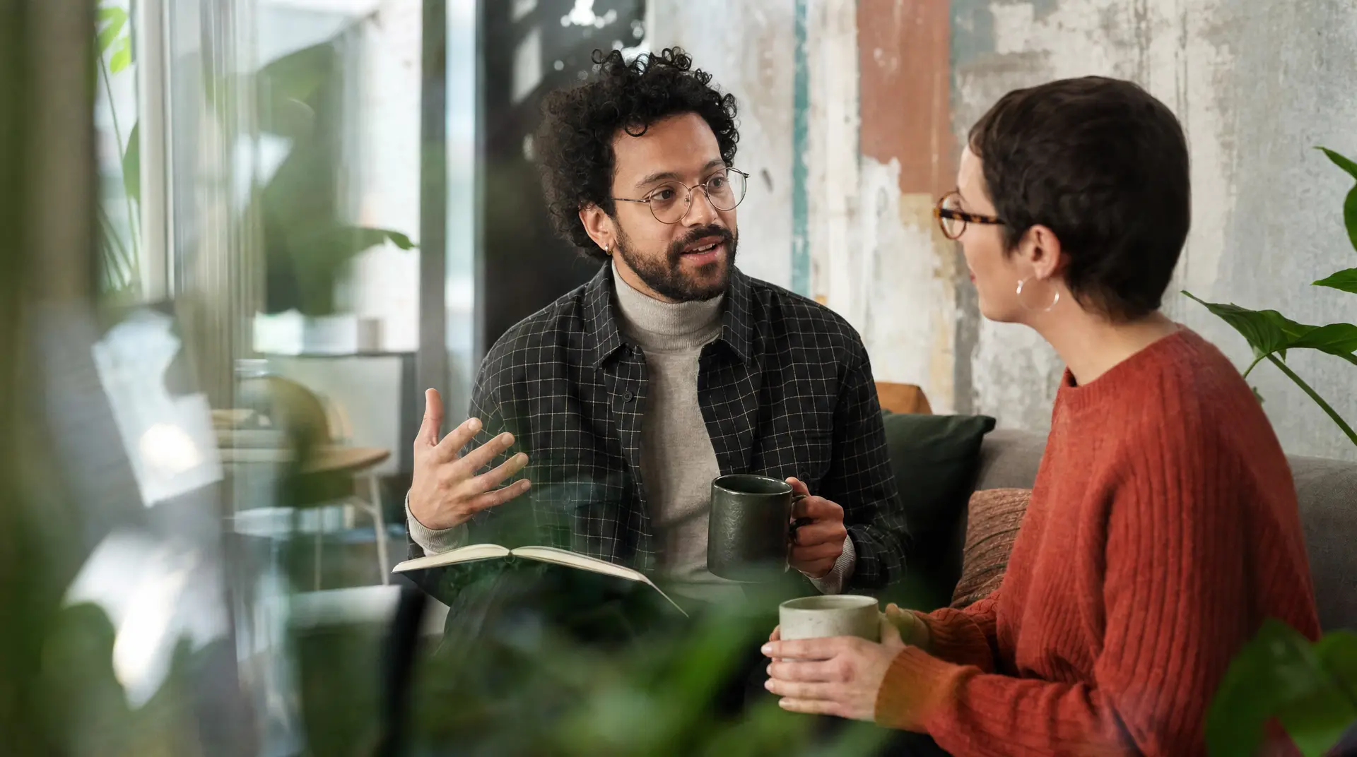 Two professionals sit in a bright office during a discussion, with one holding a mug while the other gestures, supporting improve candidate experience in recruitment.