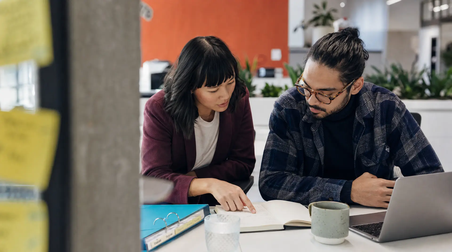 Two colleagues review notes at a desk with a laptop, reflecting recruitment onboarding work.