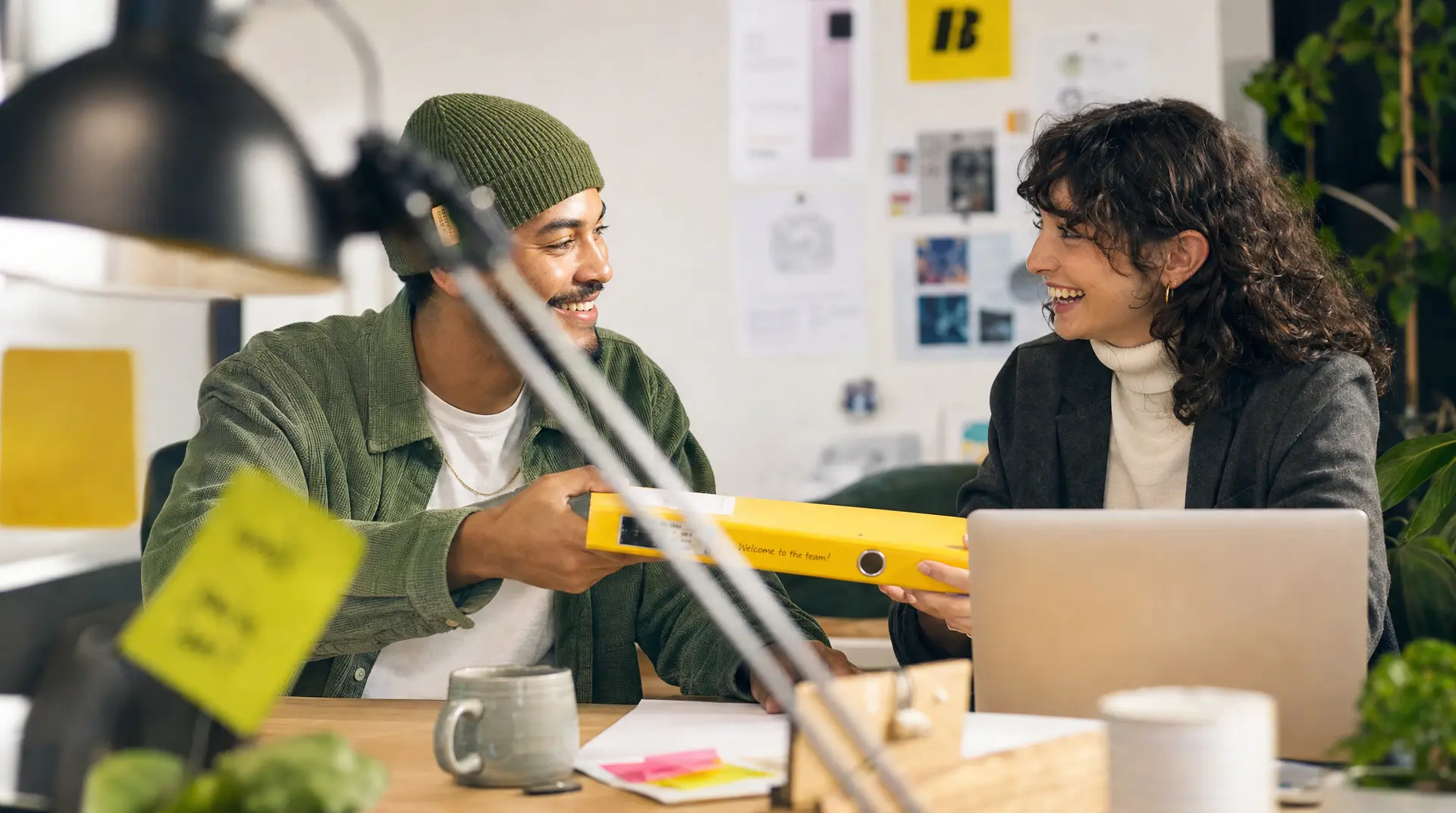Two coworkers smile and exchange a yellow binder while seated at a table with laptops, supporting the recruitment process and collaborative recruitment.