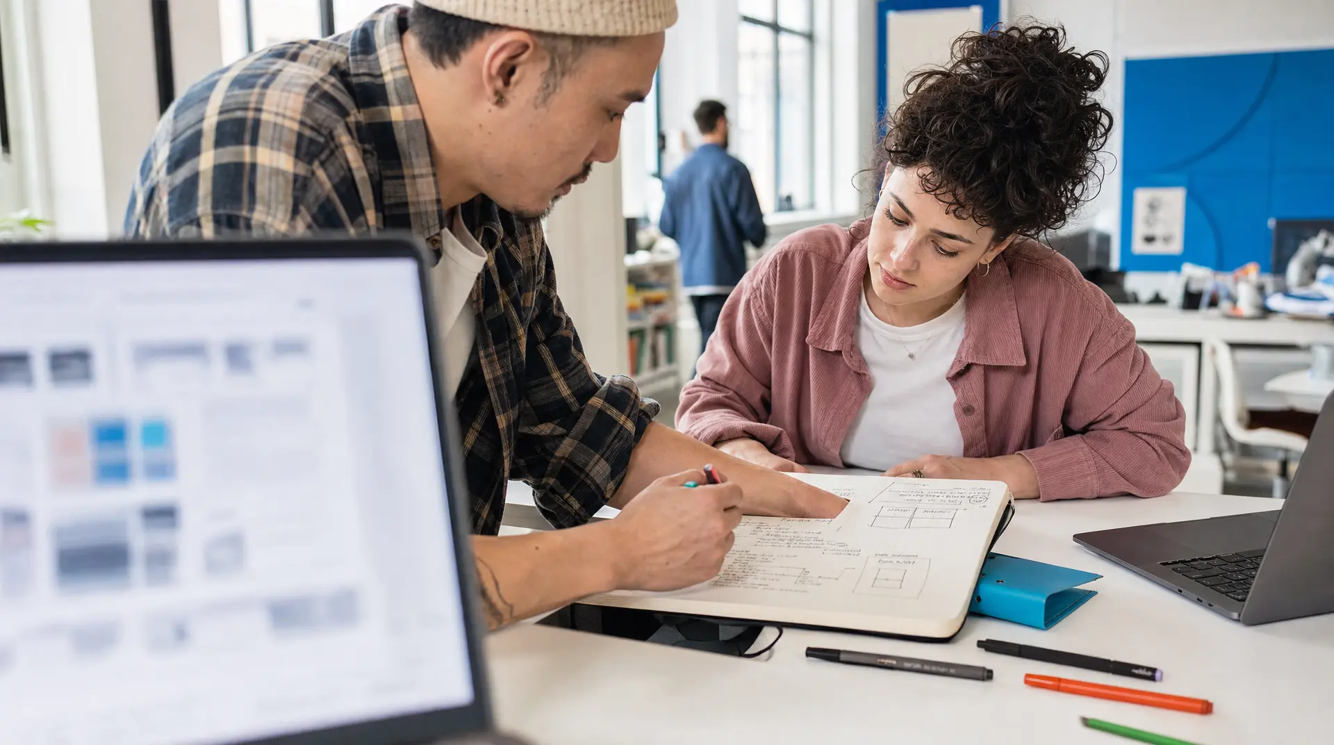 Two coworkers review notes and diagrams on a tablet and notebook at a shared desk, supporting recruitment workflow planning.
