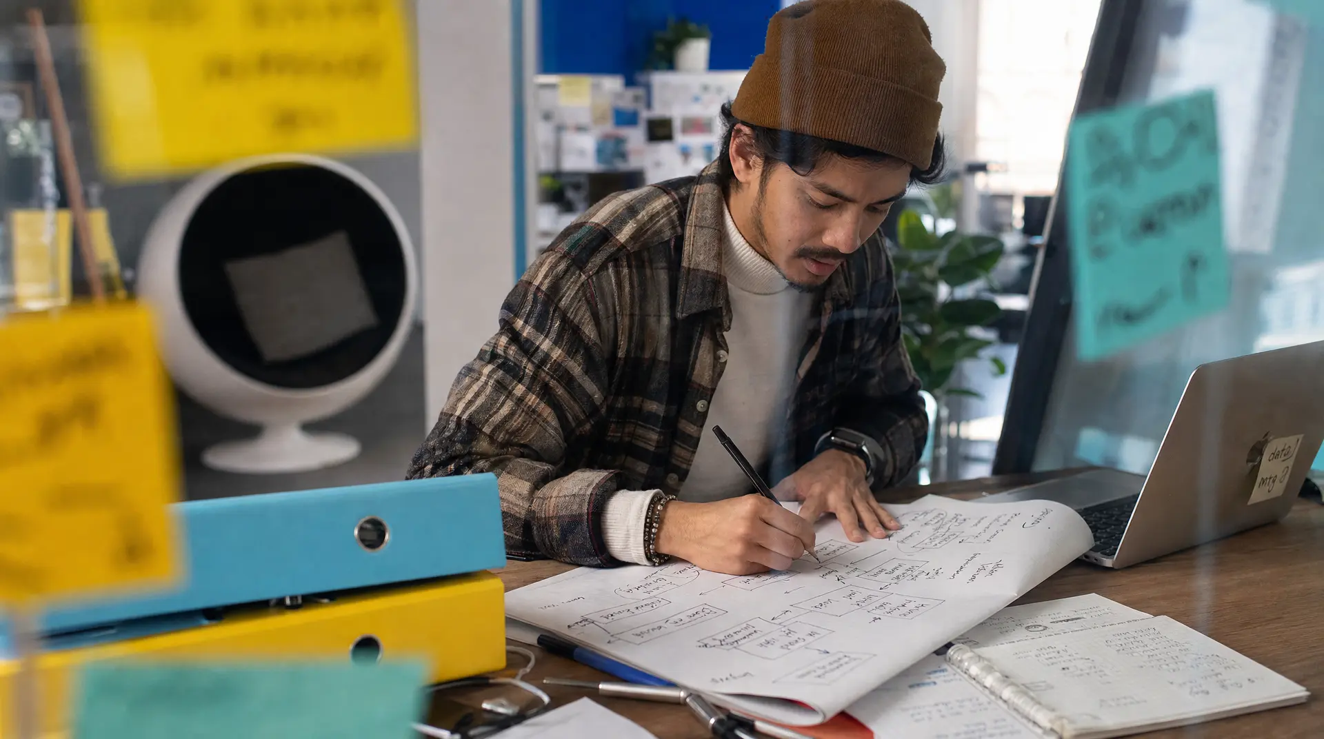 Engineer in a beanie sketches technical diagrams at a desk beside a laptop, supporting technology recruitment in a busy office environment.