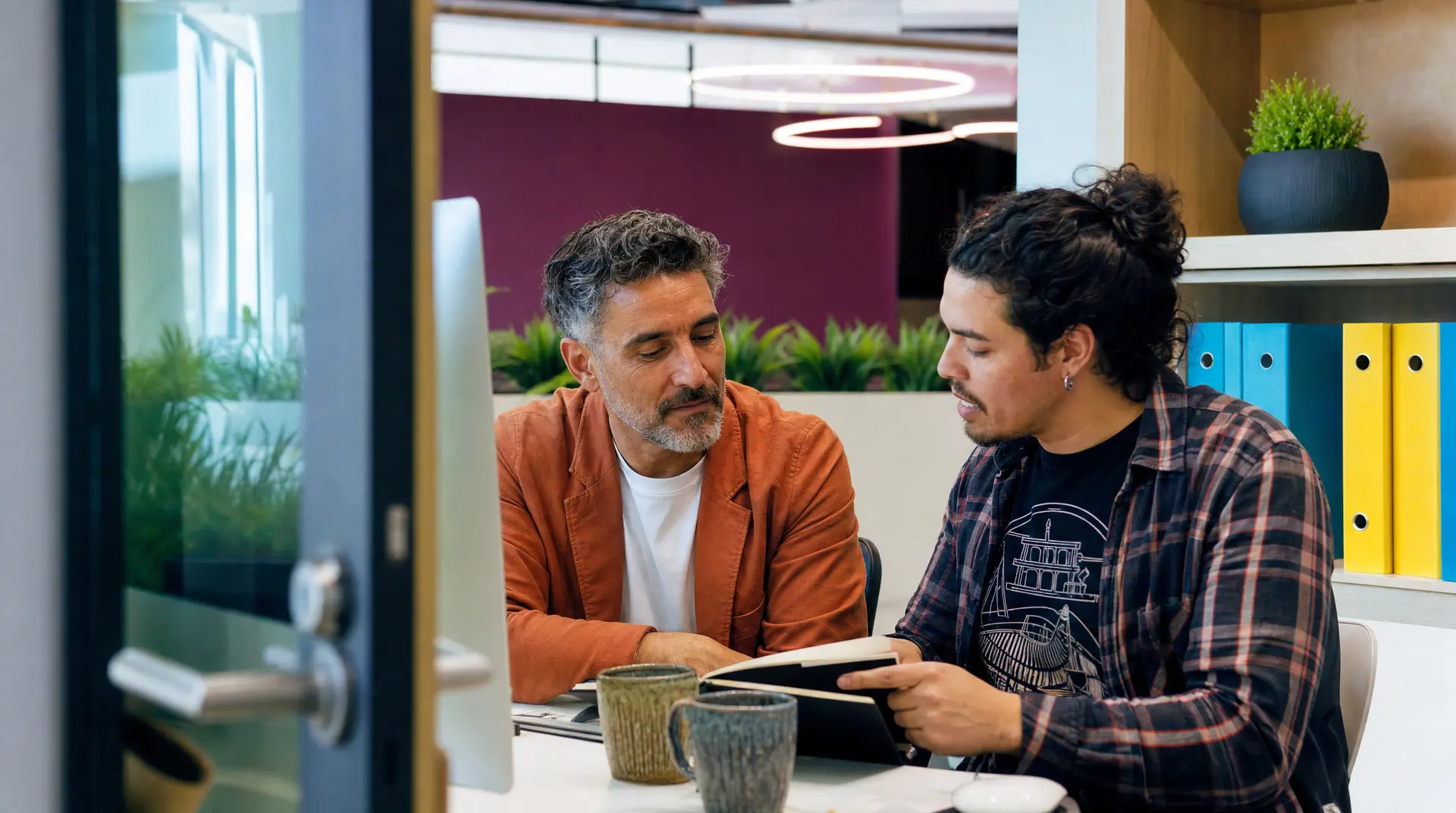 Two professionals review an open document together at a desk in a bright office, with colorful binders on shelves behind them, reflecting a recruitment process discussion.