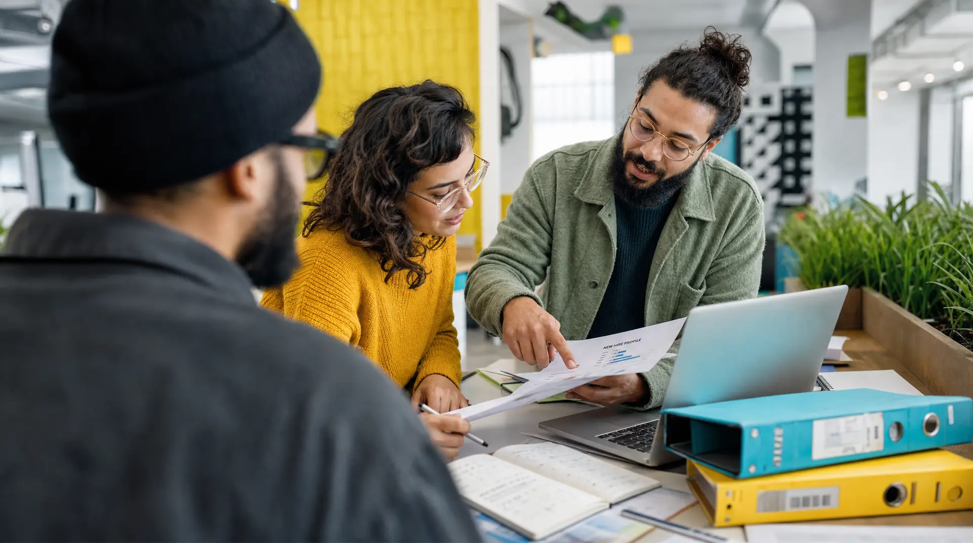 A recruiter and two candidates review printed onboarding paperwork in a bright office, aligning on next steps for talent acquisition.