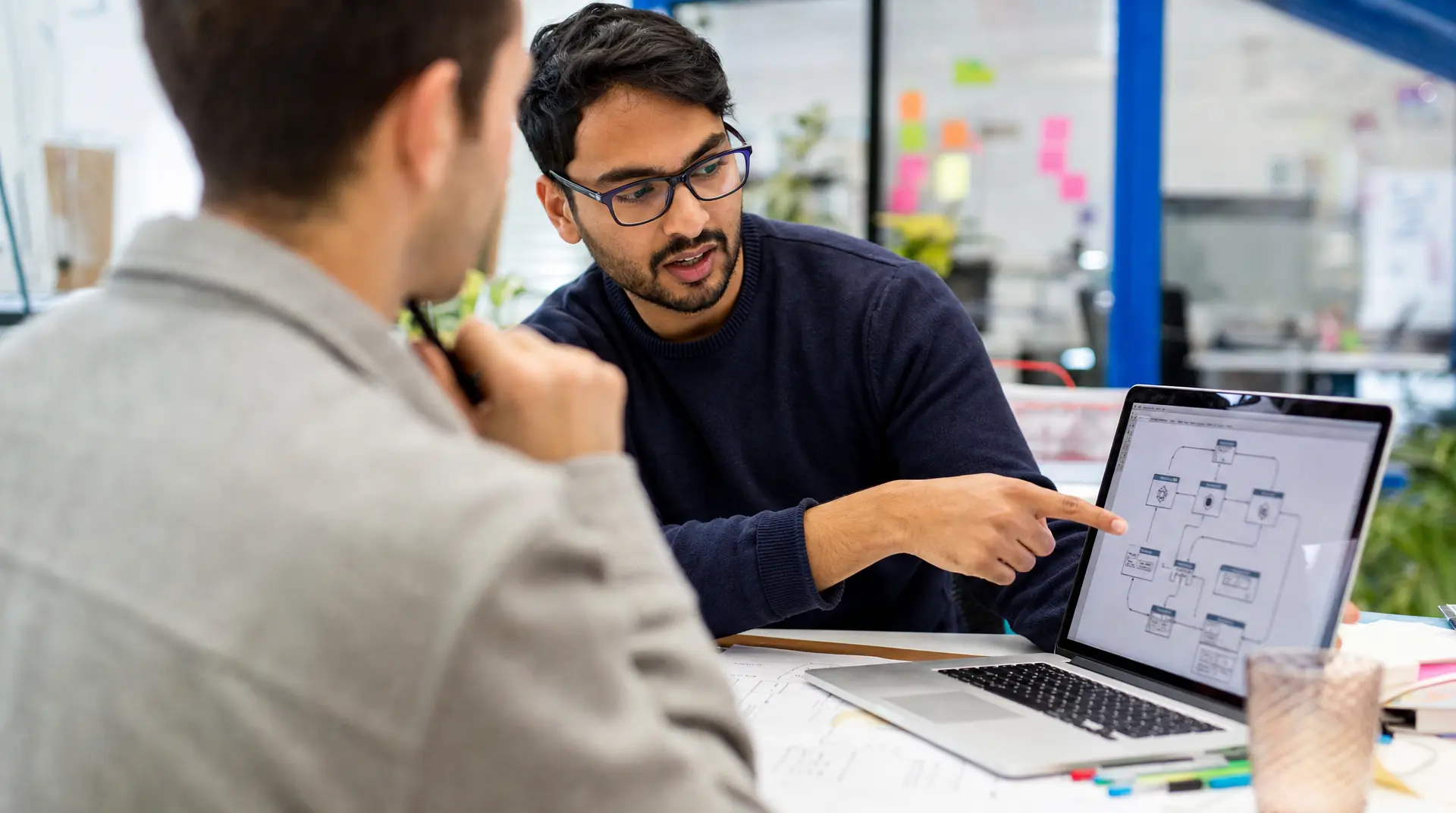 A recruiter speaks with a candidate at a desk while pointing at a laptop screen showing a system diagram, supporting technology recruitment conversations.