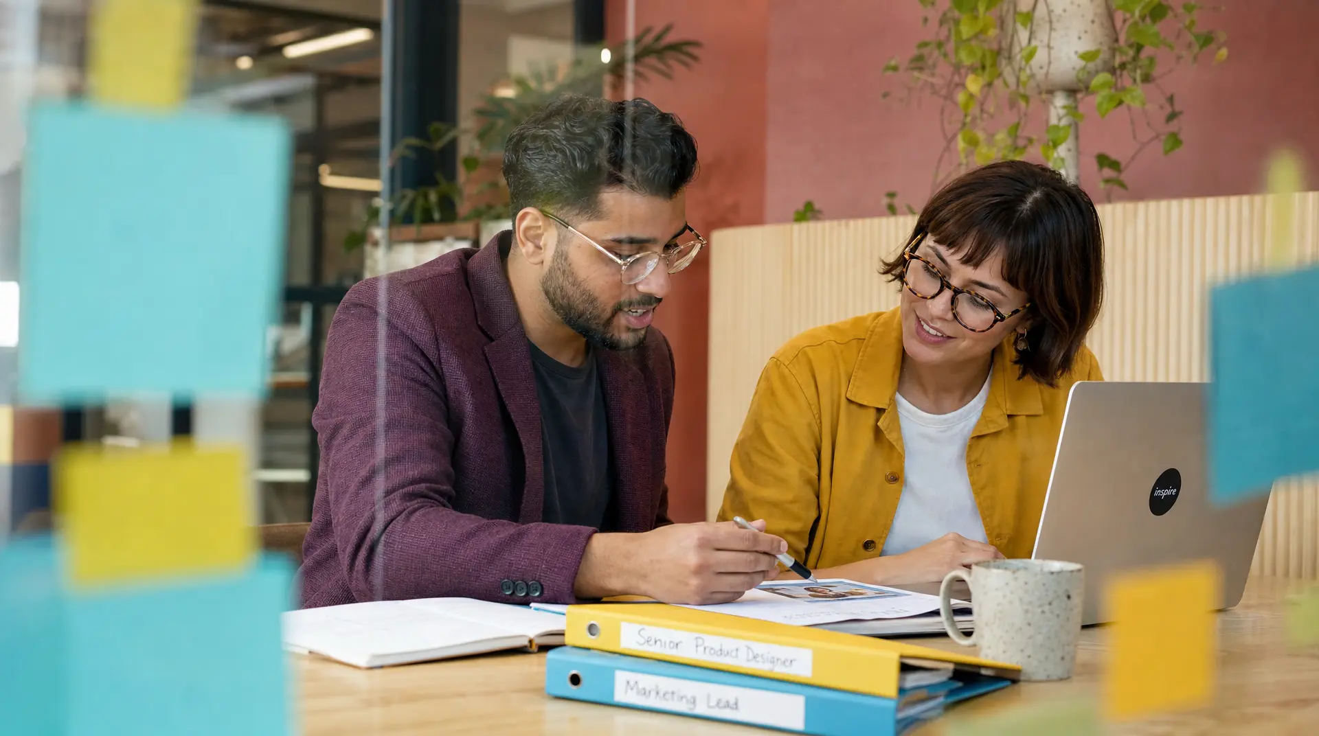 Two colleagues review product design notes at a desk with laptops open, aligning data driven recruitment decisions.