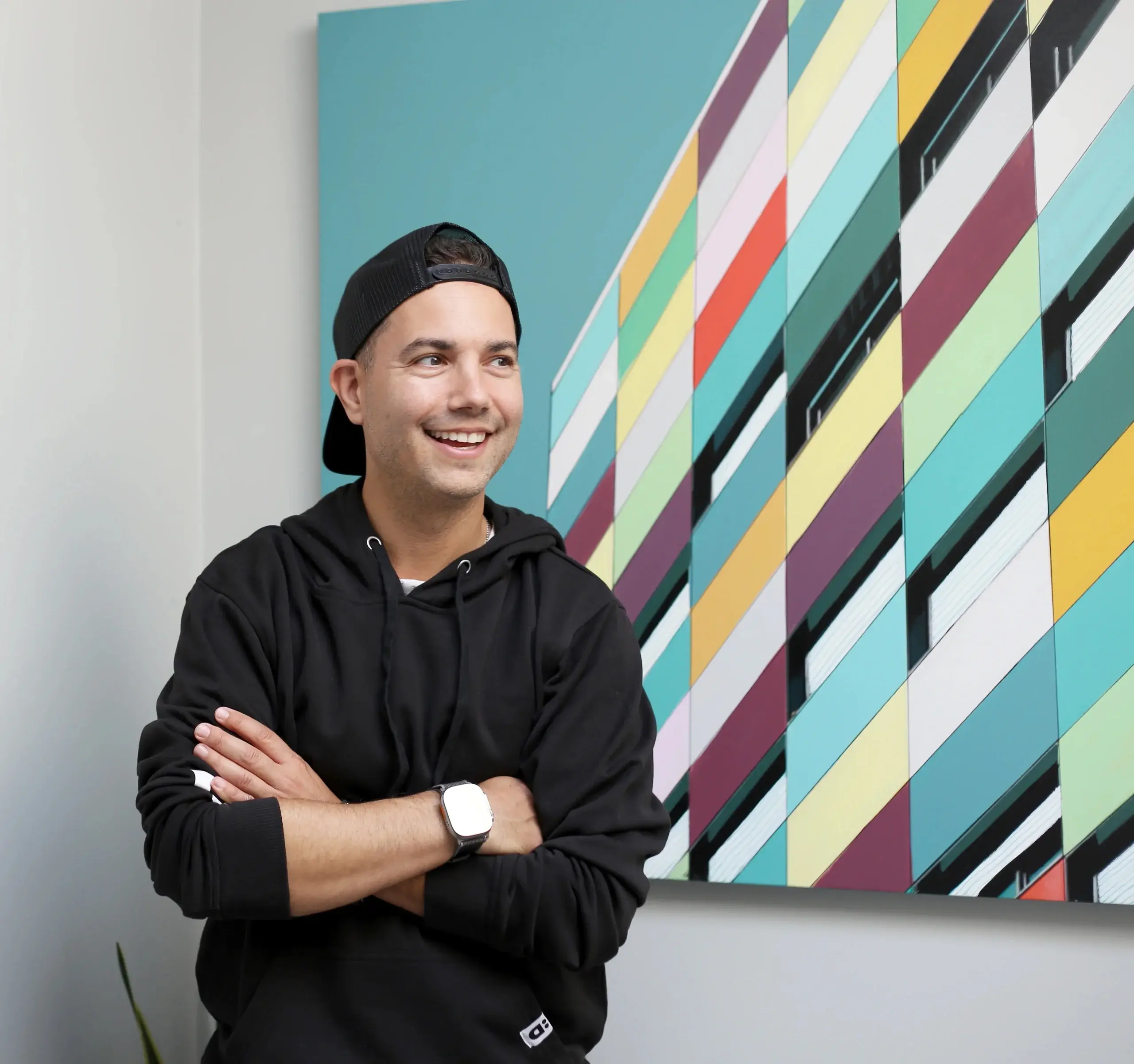 Smiling young man in a black hoodie and cap stands with arms crossed beside a colorful geometric mural, capturing gen z recruitment energy.